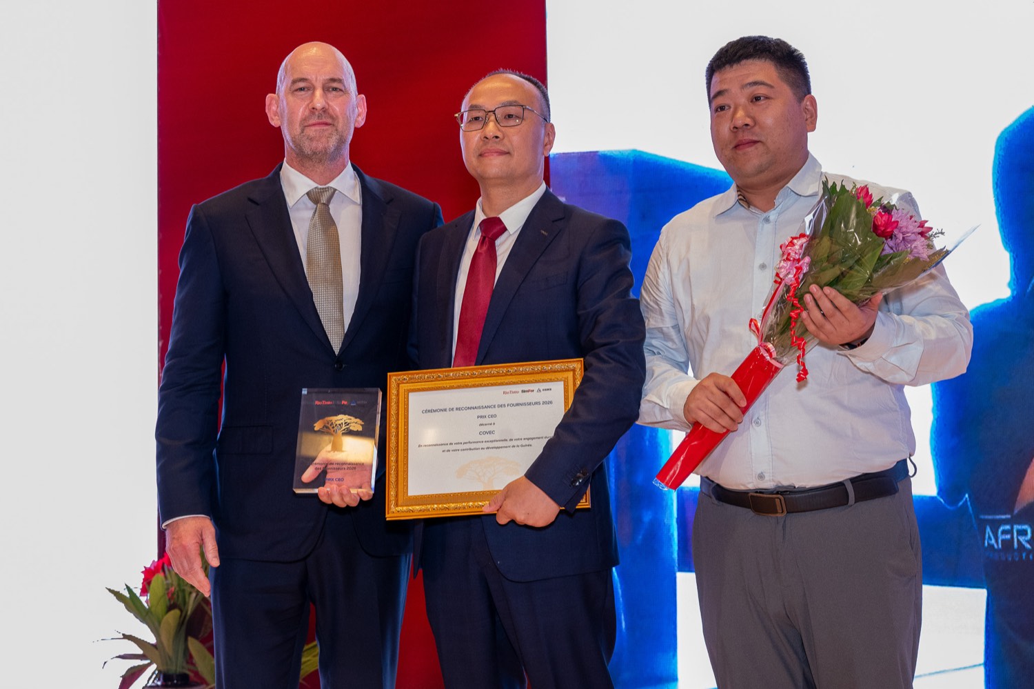 Chris Aitchison, Directeur Général de SimFer, remettant le certificat encadré du Prix du CEO à Liu Chenyu de COVEC, accompagné d'un représentant de COVEC tenant un bouquet de fleurs.